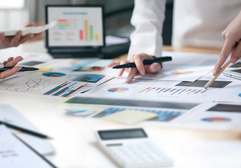 テーブル一面に広げられたグラフや資料を数人の手が指し示している